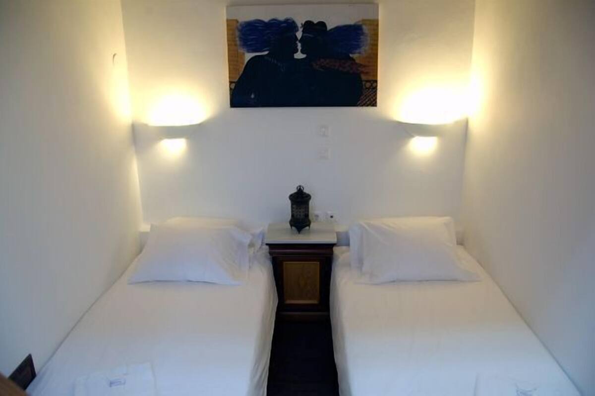 A single bed with white linens, a nightstand, a wall-mounted lamp, and a painting above the bed.