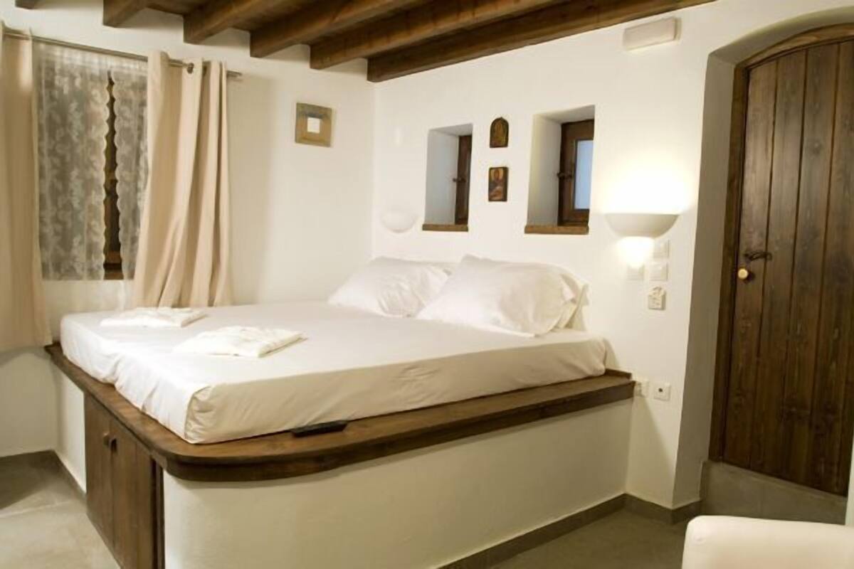 A bedroom with a wooden bed frame, a single bed with white linens, a nightstand, a wall-mounted lamp, and a wooden door.