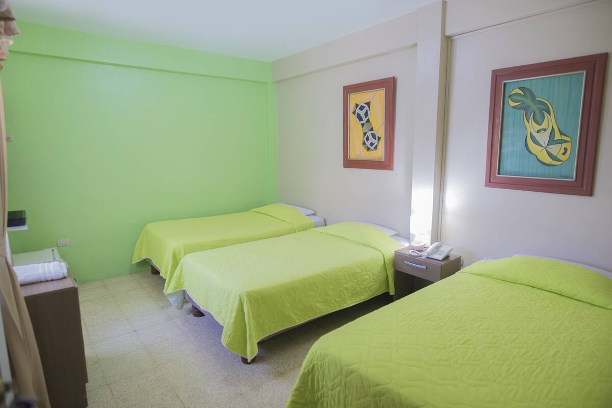 Ein Hotelzimmer mit drei Betten, einem Nachttisch mit Telefon und zwei gerahmten Kunstwerken an der Wand.