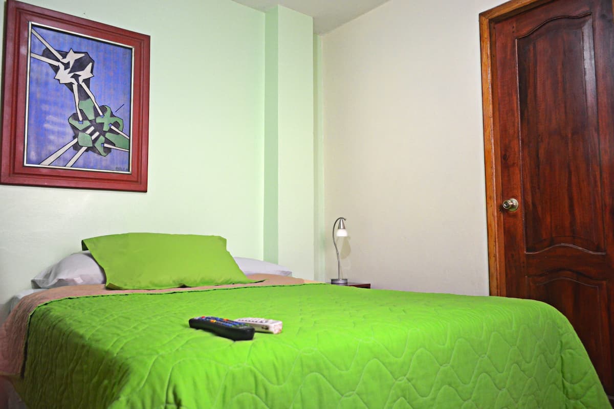 Ein Schlafzimmer mit grüner Bettdecke, einem Bild an der Wand, einer Holztür und einem Nachttisch mit Lampe.