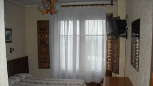 Une chambre avec deux lits, une commode en bois, un téléviseur fixé au mur et une fenêtre avec des rideaux légers.