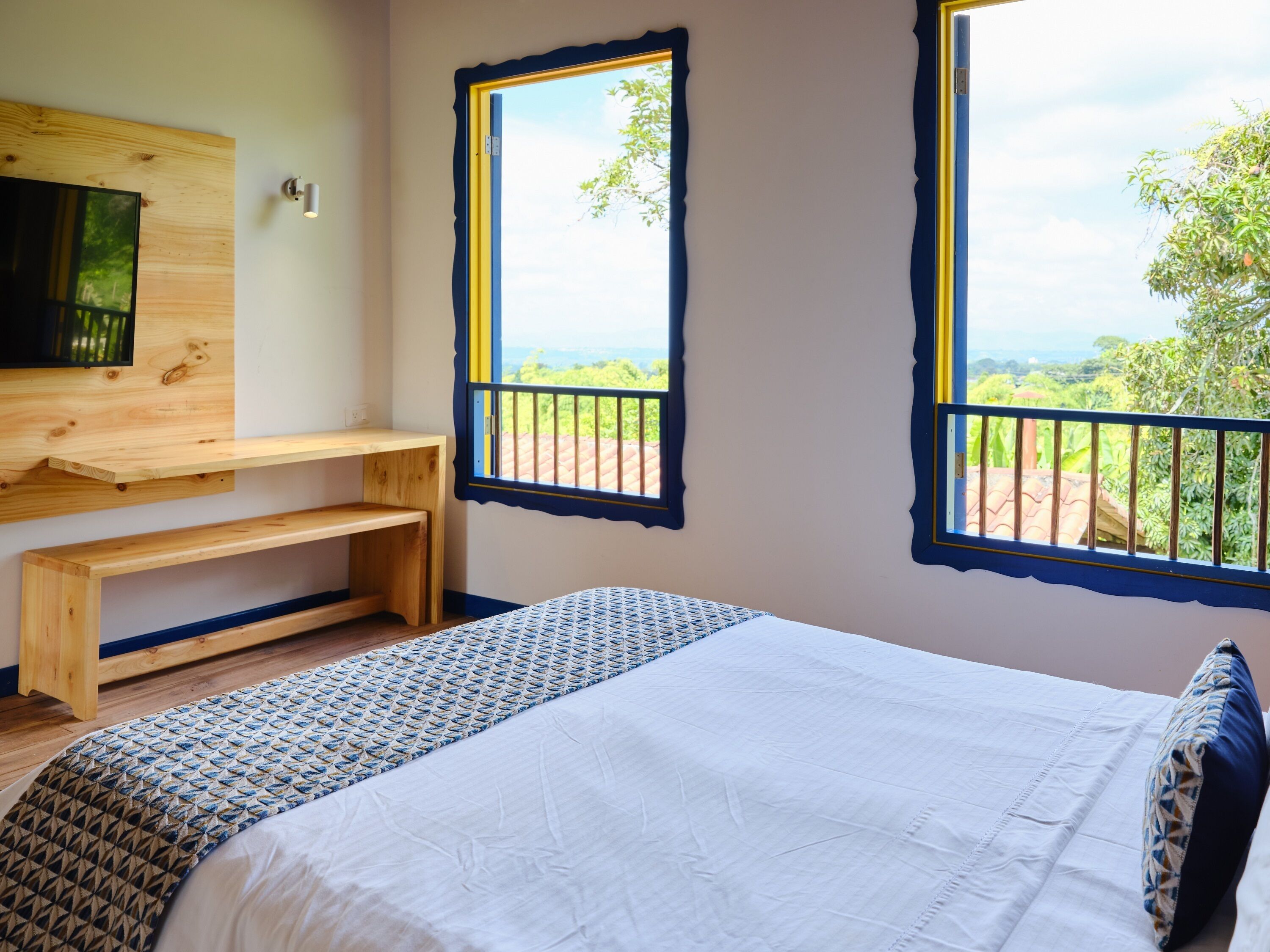 A bedroom with a bed, a wooden desk, and two windows with a view of greenery.