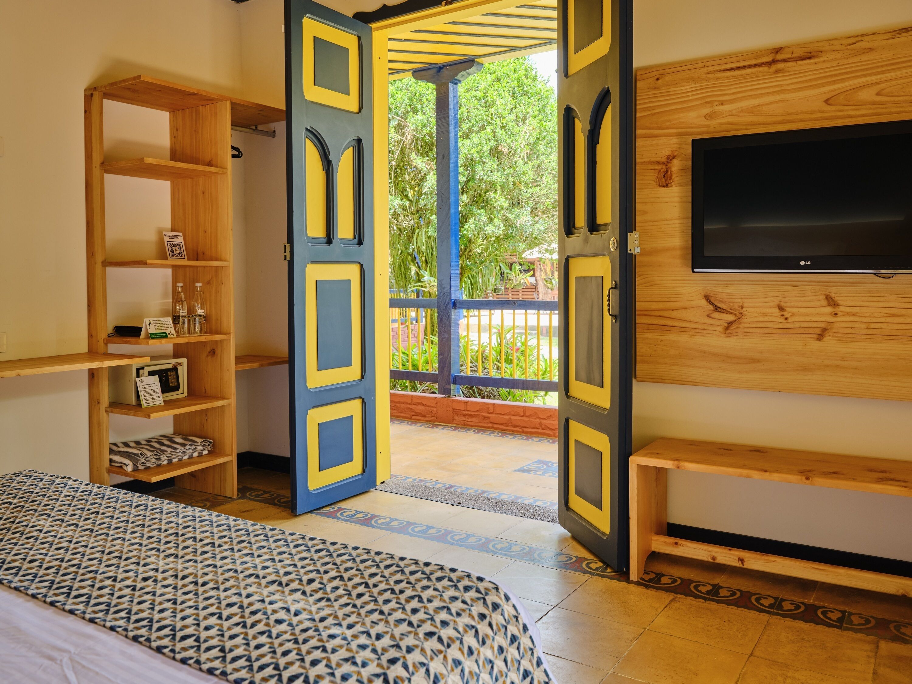 Une chambre avec un lit, un téléviseur fixé au mur, une étagère en bois avec divers objets, et une porte ouverte donnant sur un espace extérieur.