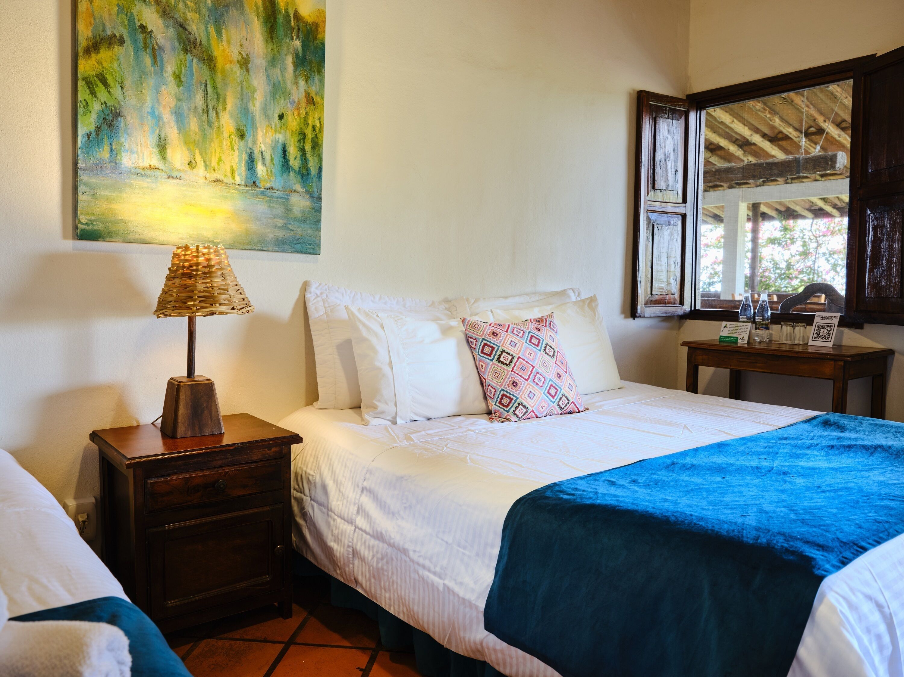 Un dormitorio con cama, una mesita de noche con lámpara, un cuadro en la pared y una ventana con vistas al exterior.