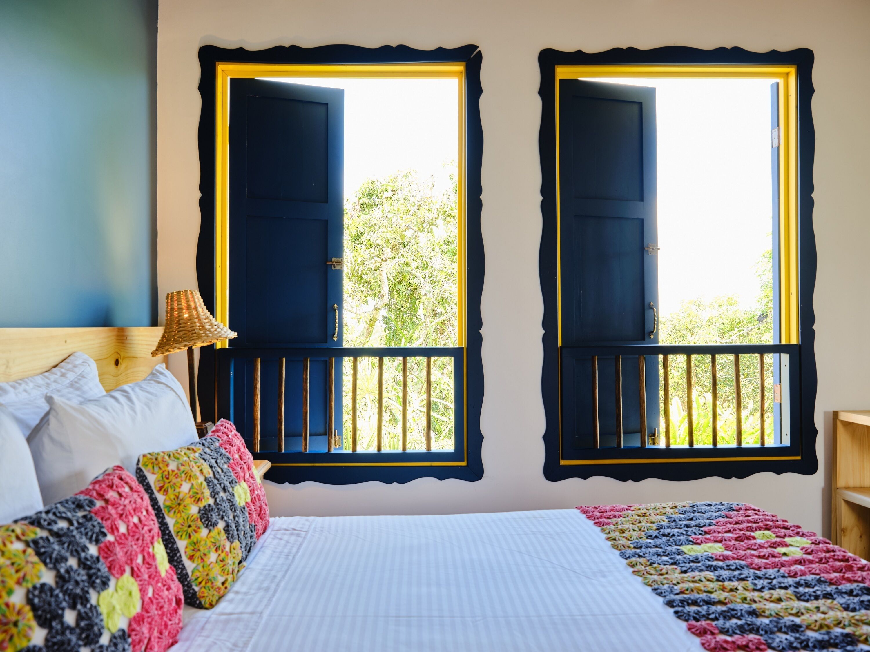 A bedroom with a bed, pillows, a lamp, and two windows with black shutters.