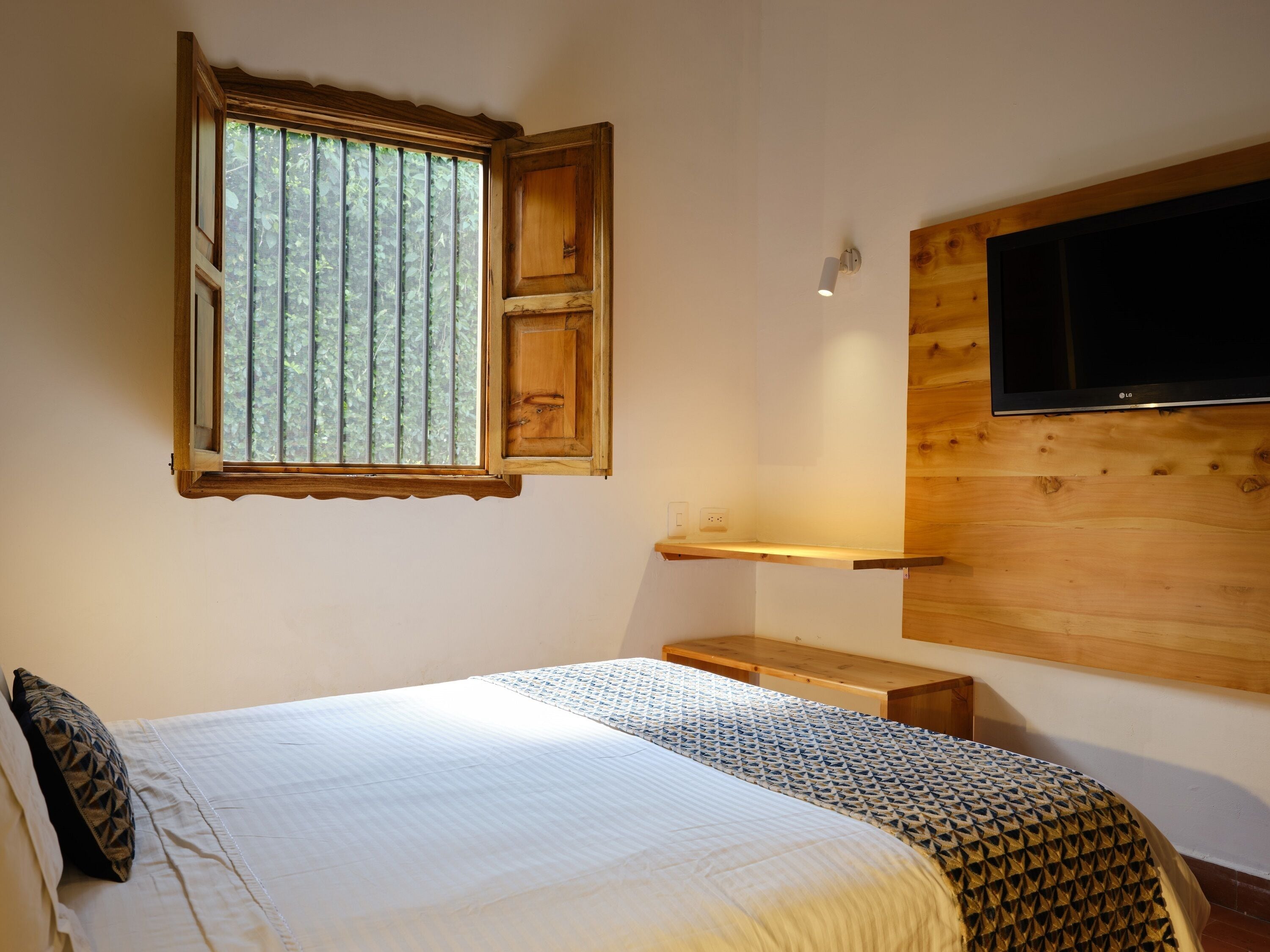 Une chambre à coucher avec un lit, une fenêtre avec des stores, une étagère en bois et un téléviseur fixé au mur.