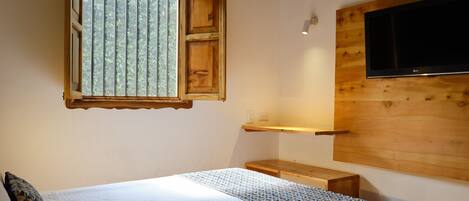 Une chambre à coucher avec un lit, une fenêtre avec des stores, une étagère en bois et un téléviseur fixé au mur.