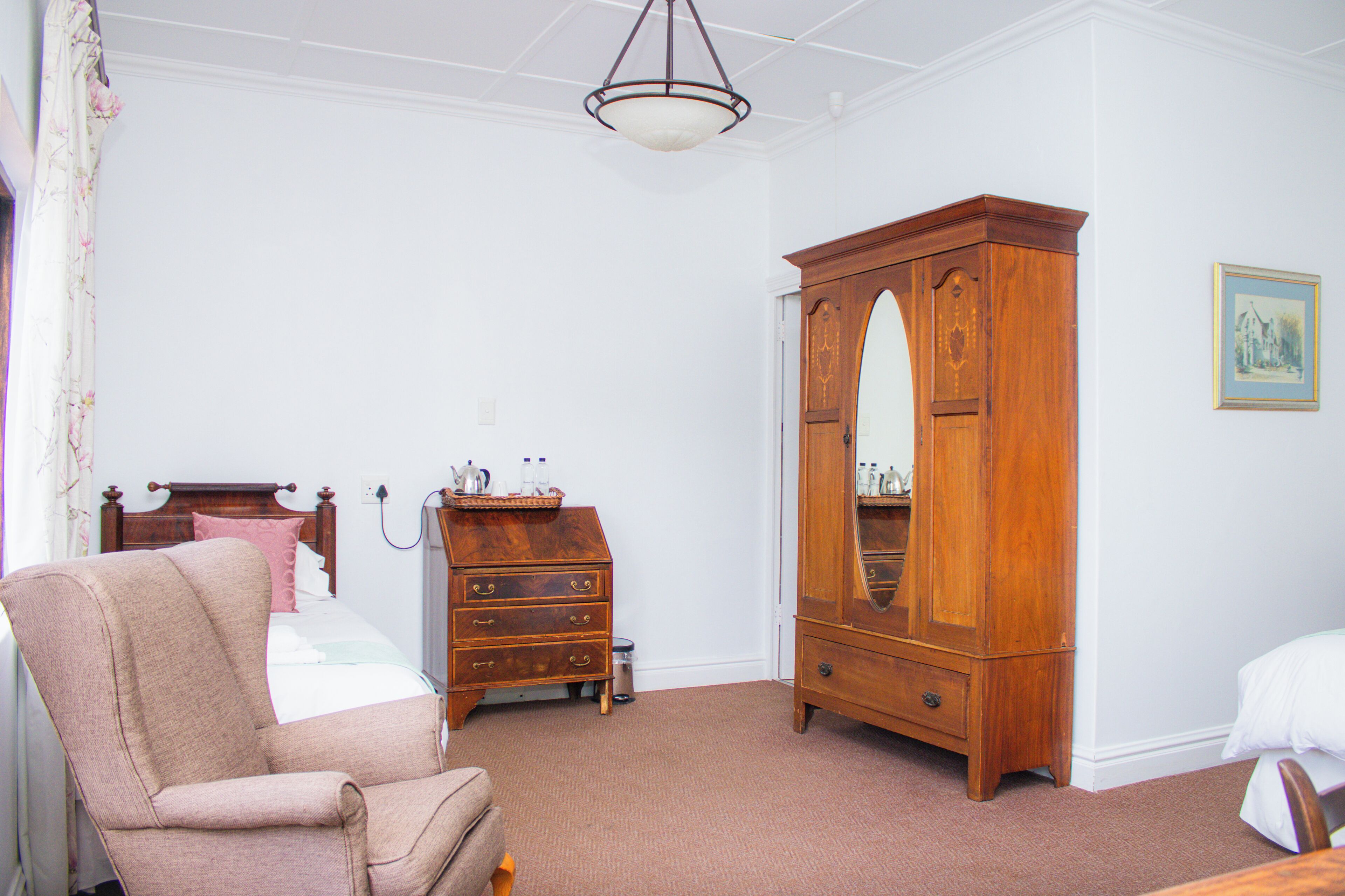 A room with a wooden armoire, a chair, a bed with a headboard, and a painting on the wall.