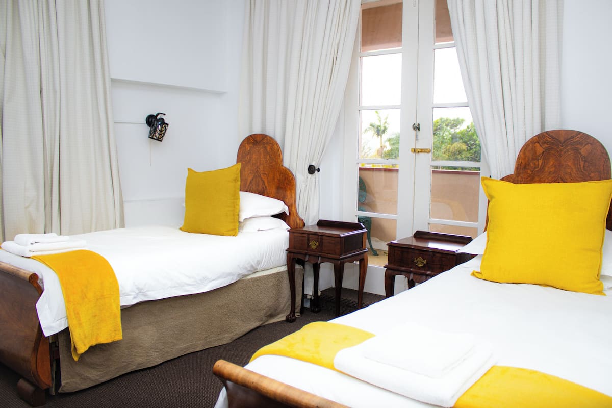 A hotel room with two beds, each with a wooden headboard and yellow pillows. There is a small wooden nightstand between the beds, and a window with curtains.