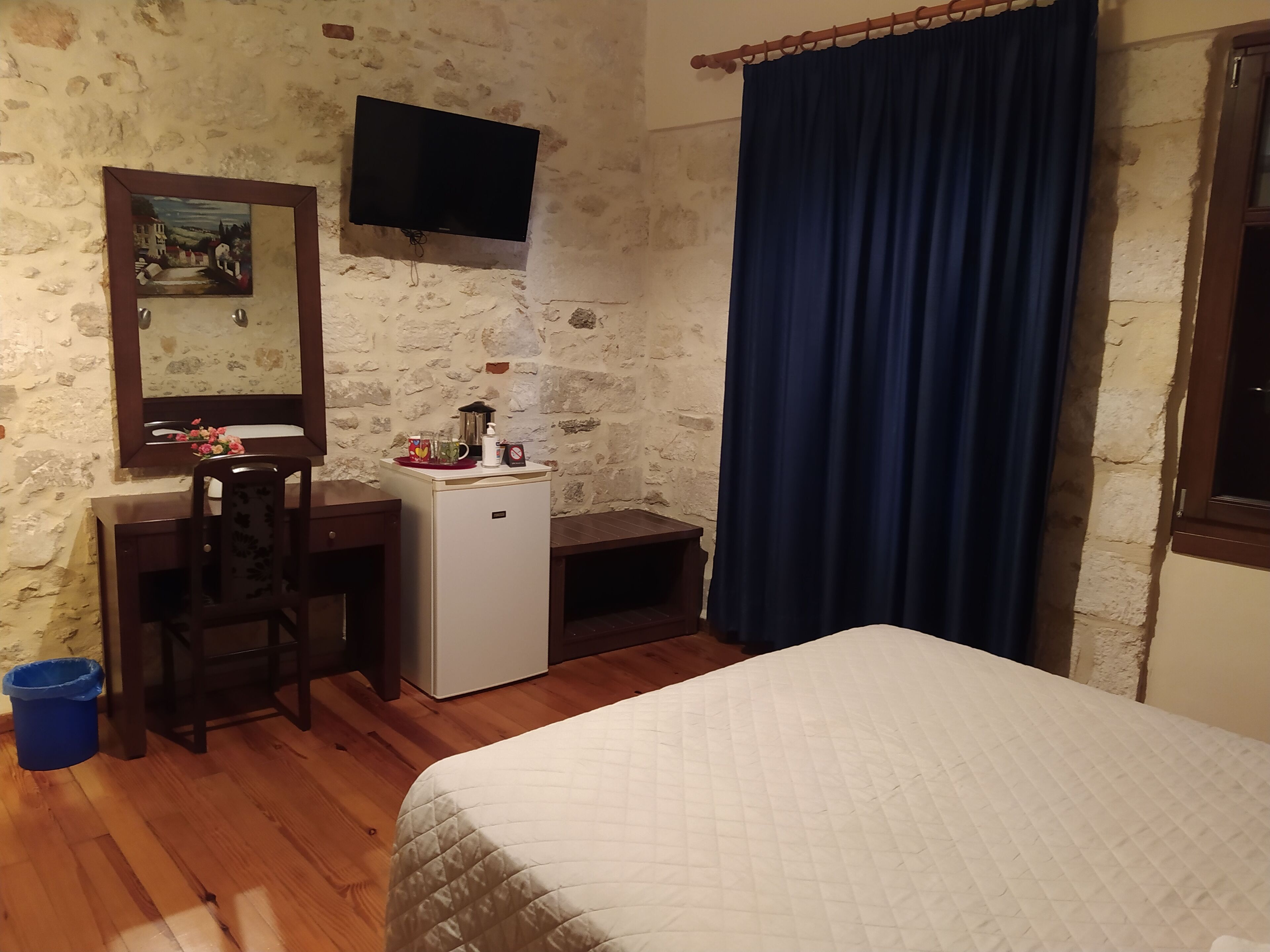 Une chambre à coucher comprenant un lit, un bureau, un petit réfrigérateur, un téléviseur fixé au mur et un rideau bleu.