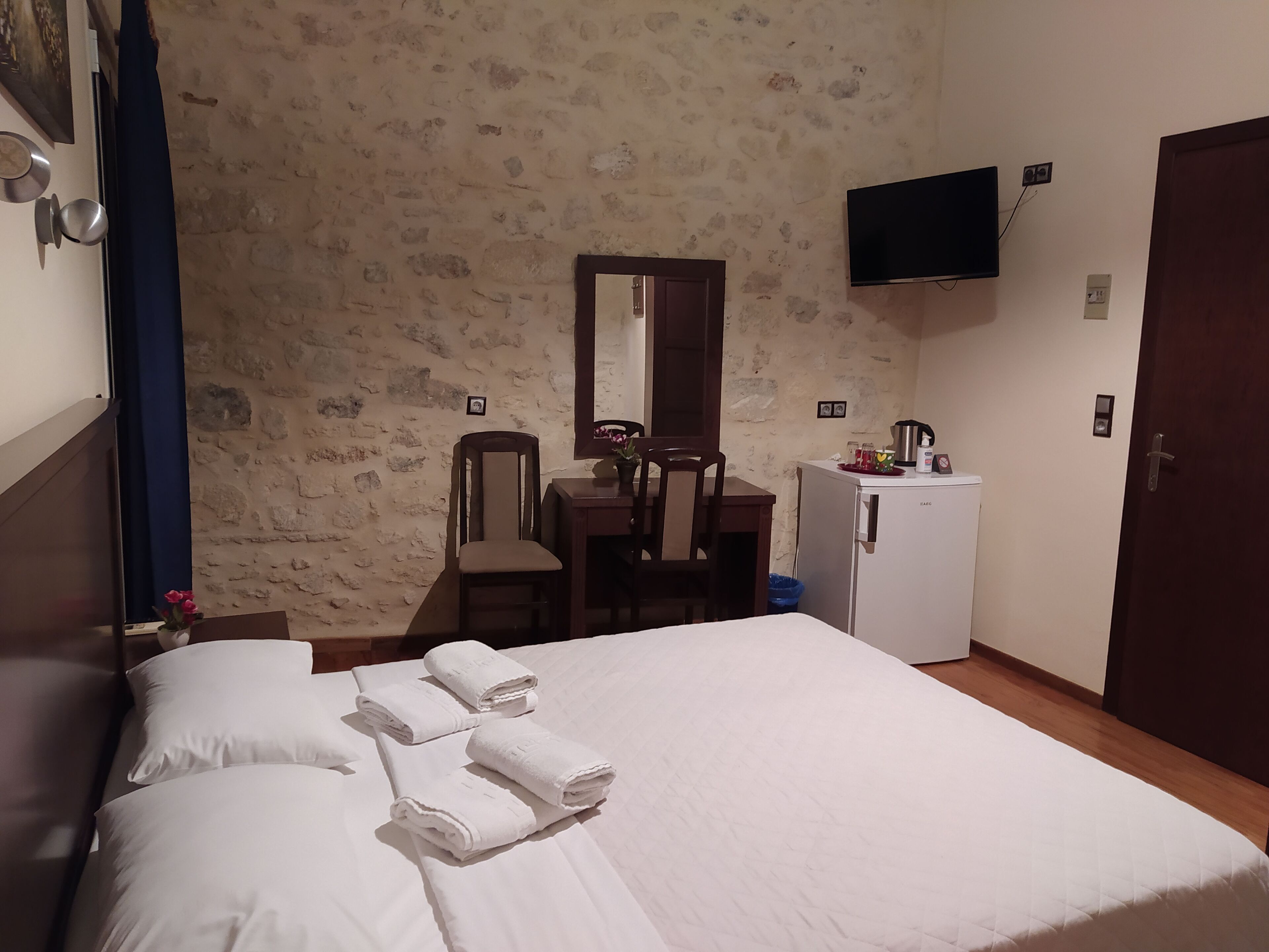 Una habitación de hotel con cama, escritorio con silla, refrigerador pequeño, televisor montado en la pared y una puerta.