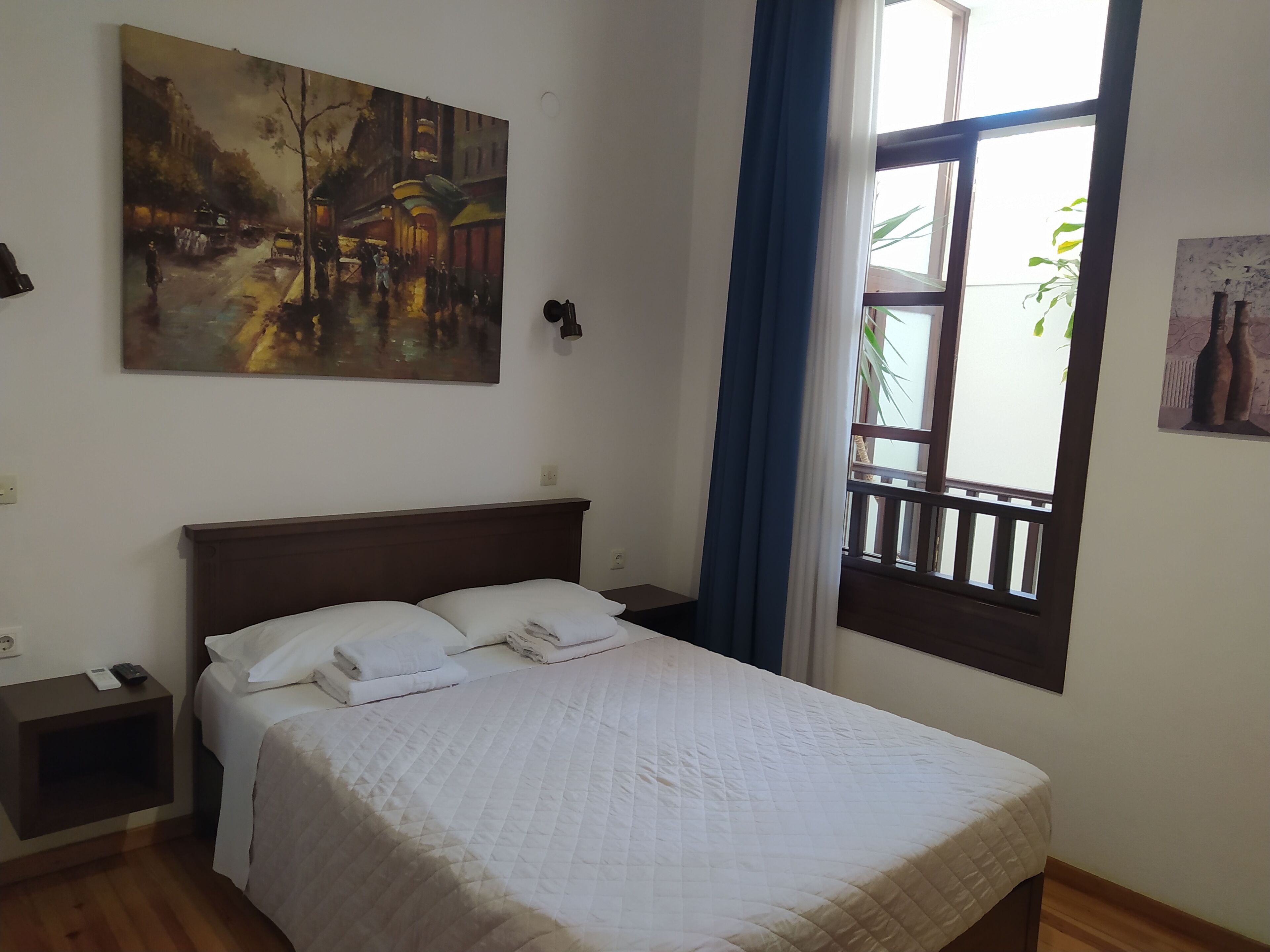 A bedroom with a bed, a nightstand, a painting, a window with a view, and a curtain.