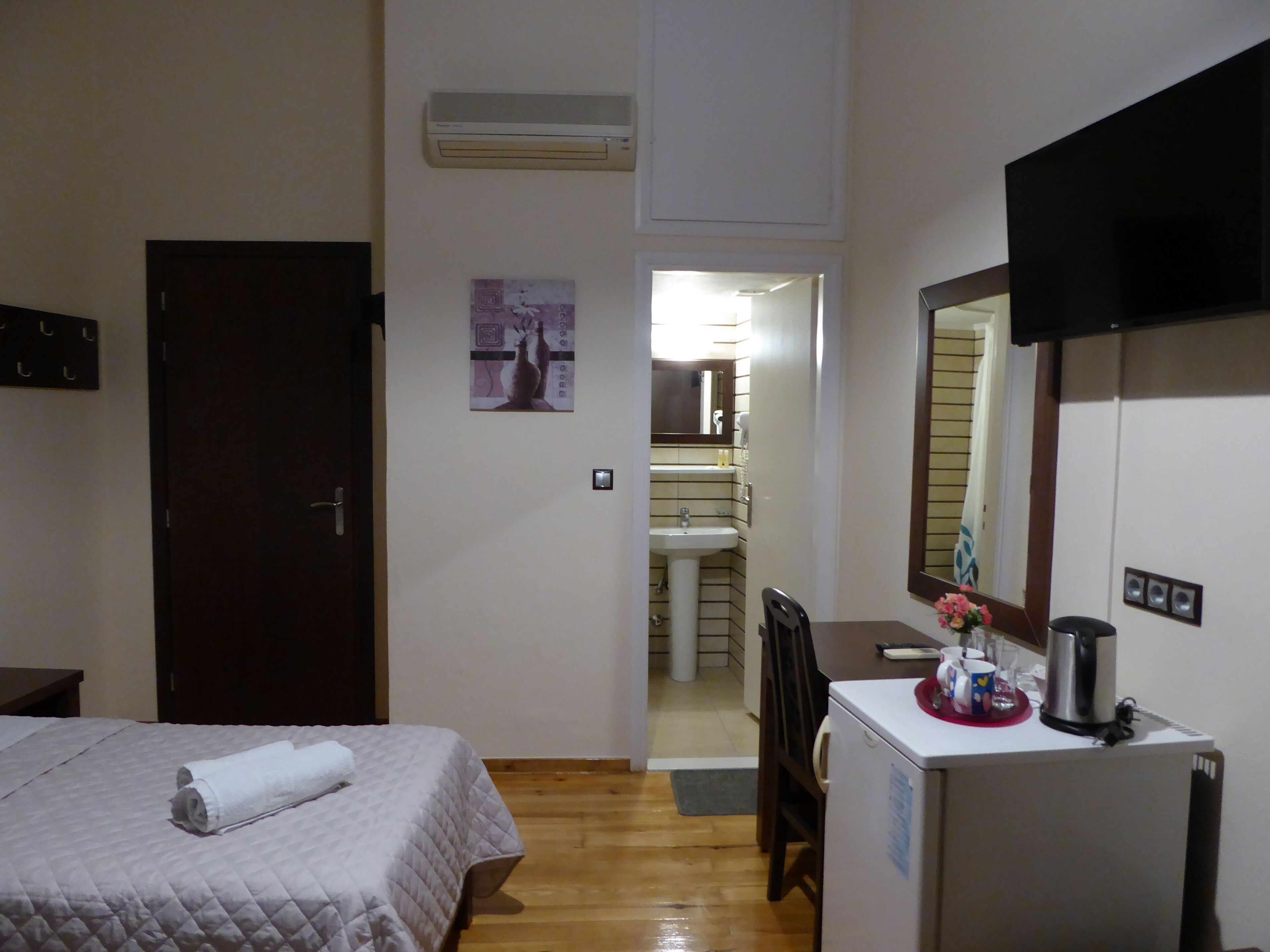 A hotel room with a bed, a desk with a refrigerator, a mirror, and a bathroom visible through an open door.