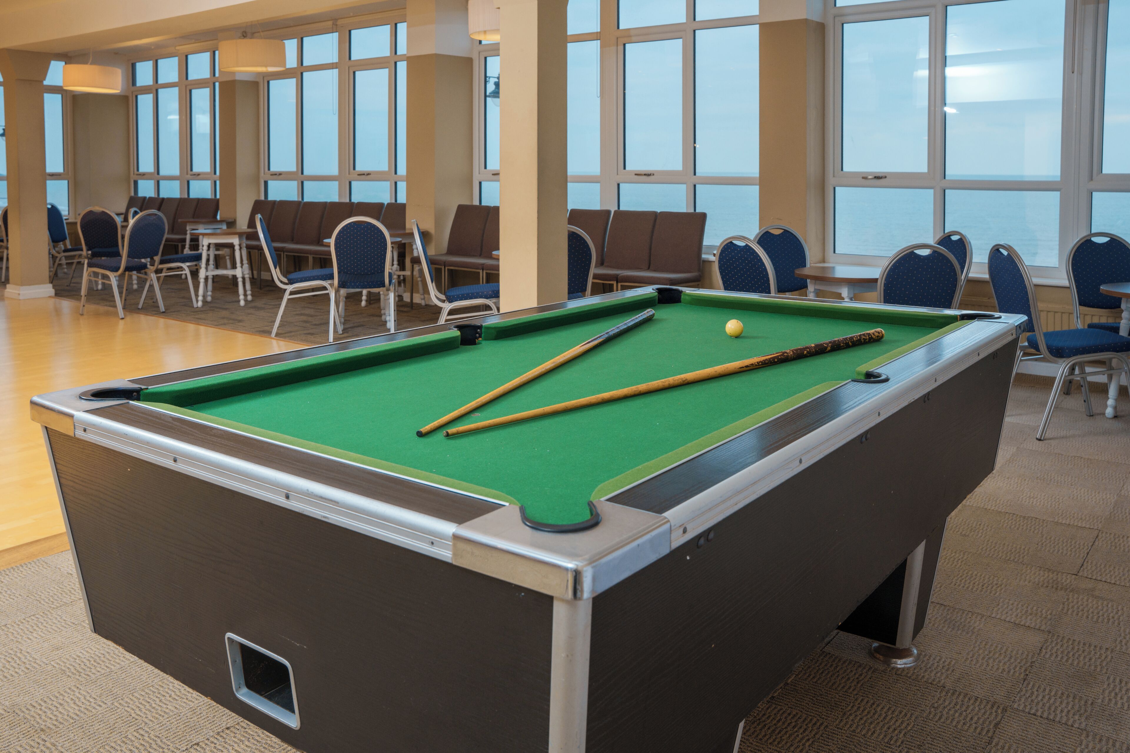 Ein Billardtisch in einem Raum mit großen Fenstern, Stühlen und Blick aufs Meer.
