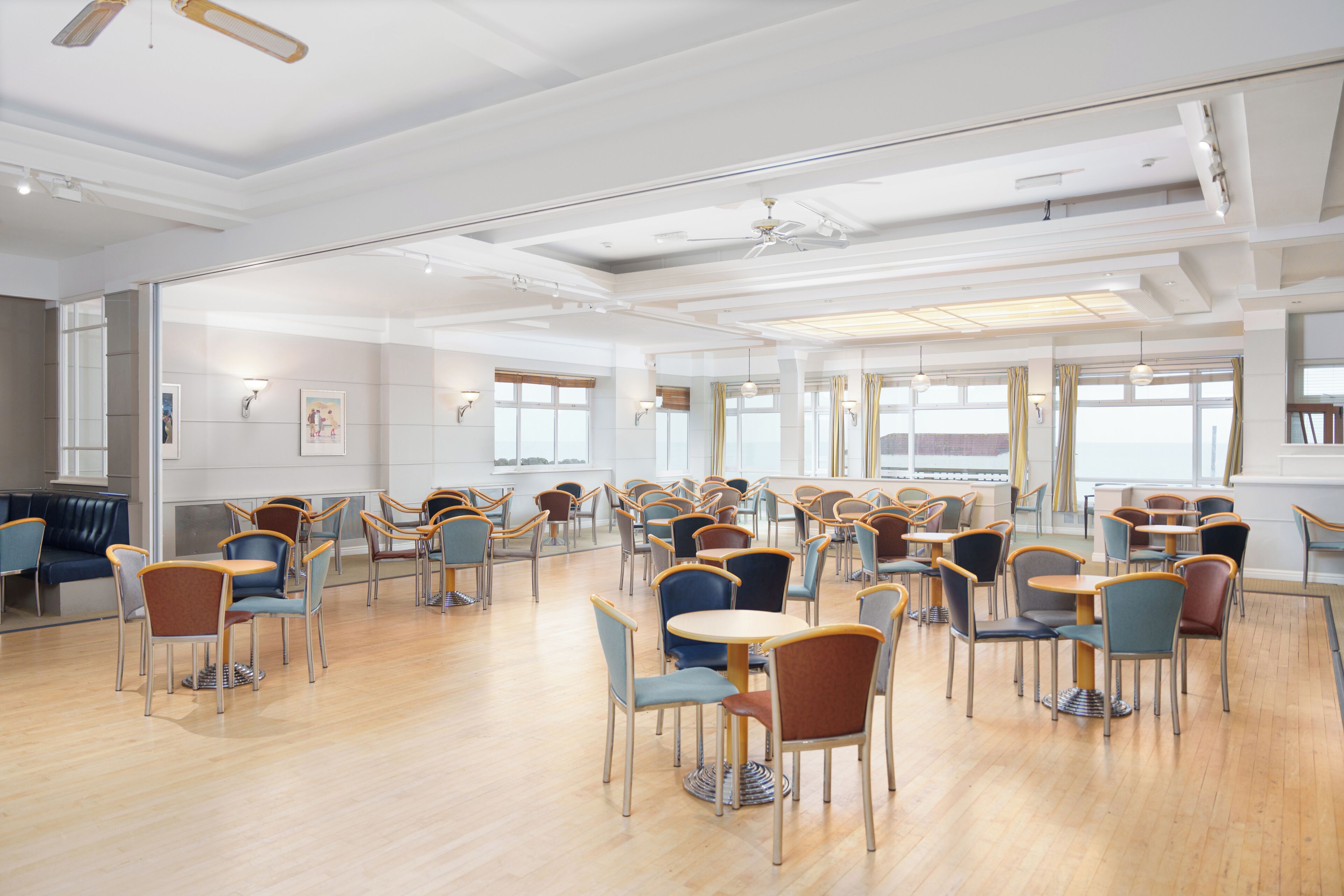 A spacious room with multiple tables and chairs, large windows, and a ceiling fan.