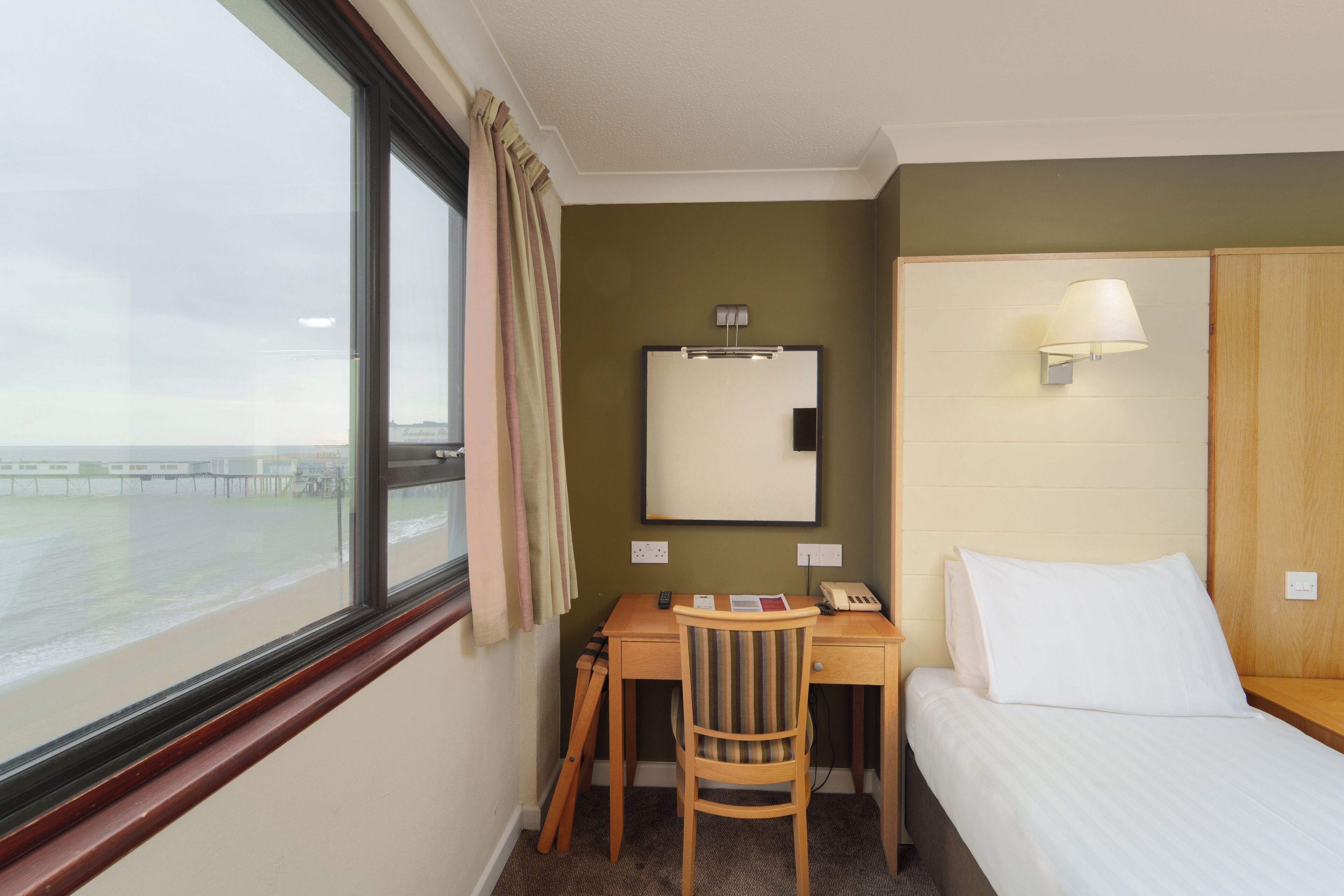 A hotel room with a desk, chair, bed, and a view of the beach and pier through a window.