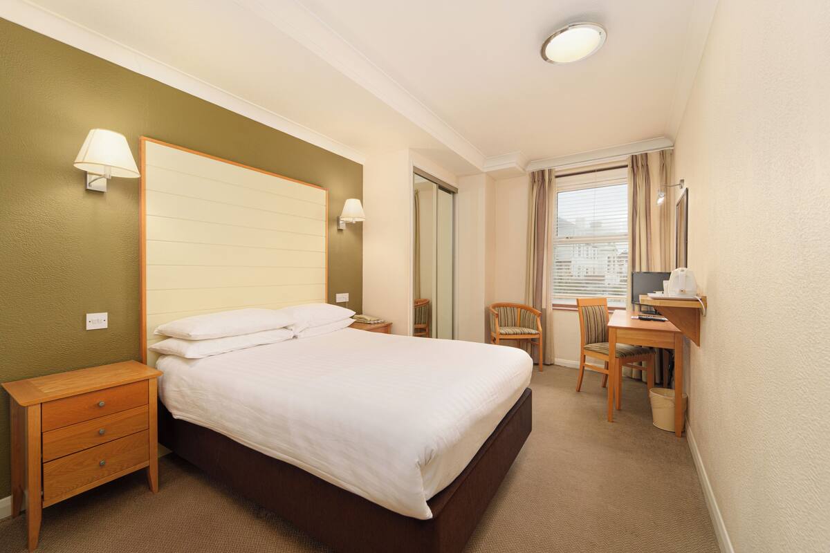 A hotel room with a large bed, a desk with a chair, a nightstand, and a window with curtains.