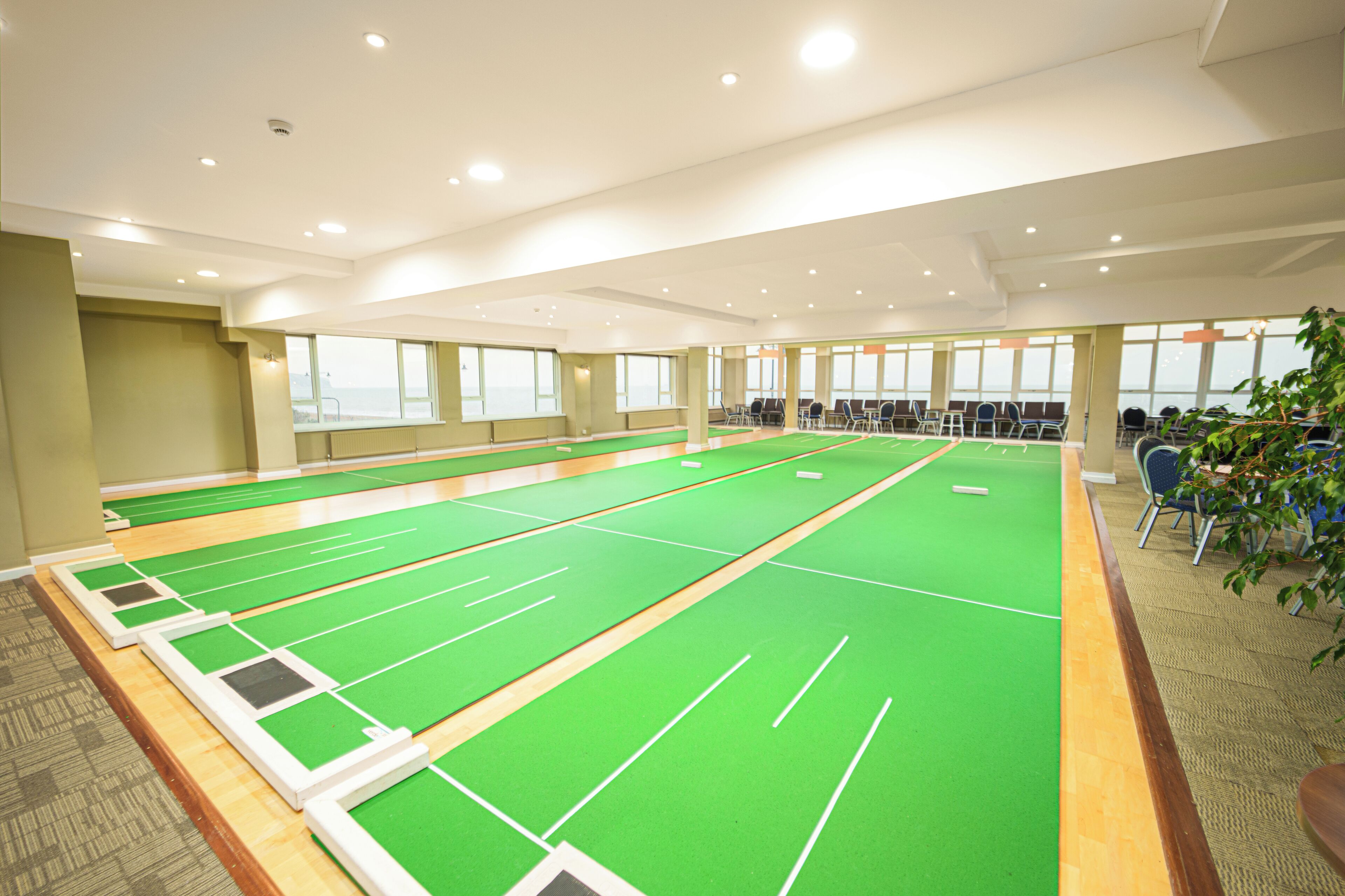 A large indoor space with multiple green shuffleboard tables and seating areas.