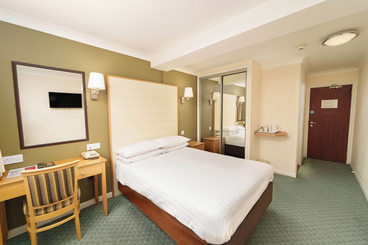 A hotel room with a bed, a desk, a chair, a mirror, a nightstand, and a door.