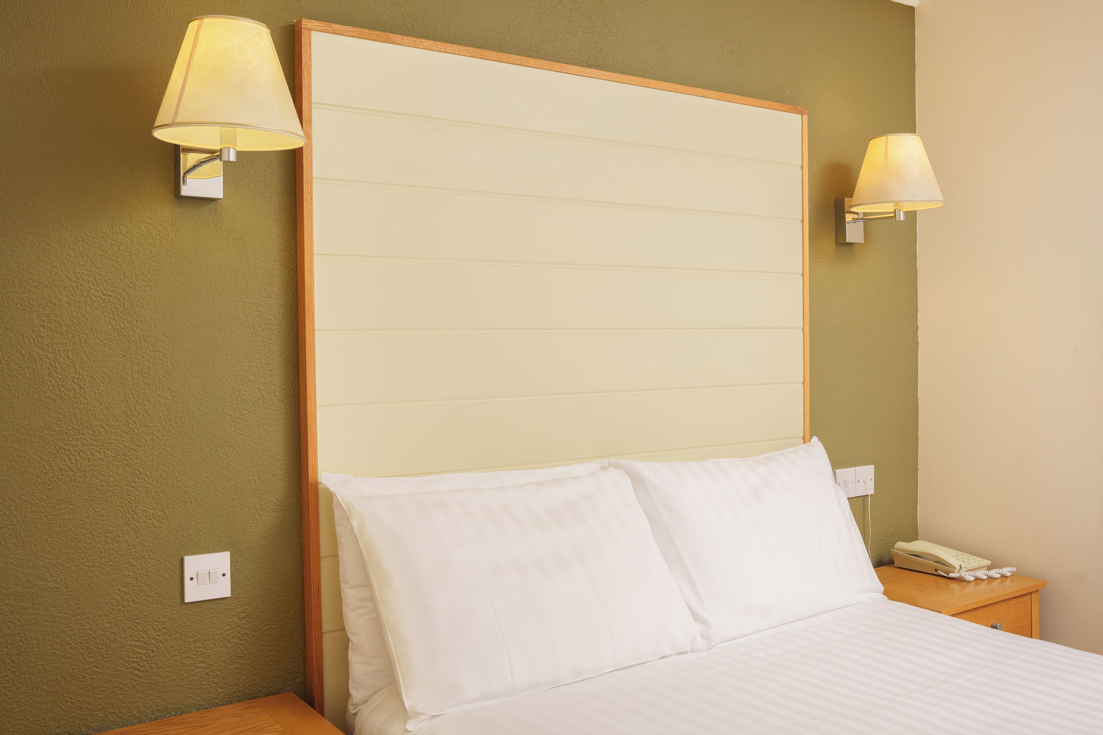 A hotel room with a large bed, two bedside lamps, a wooden headboard, a nightstand with a telephone, and a wall socket.