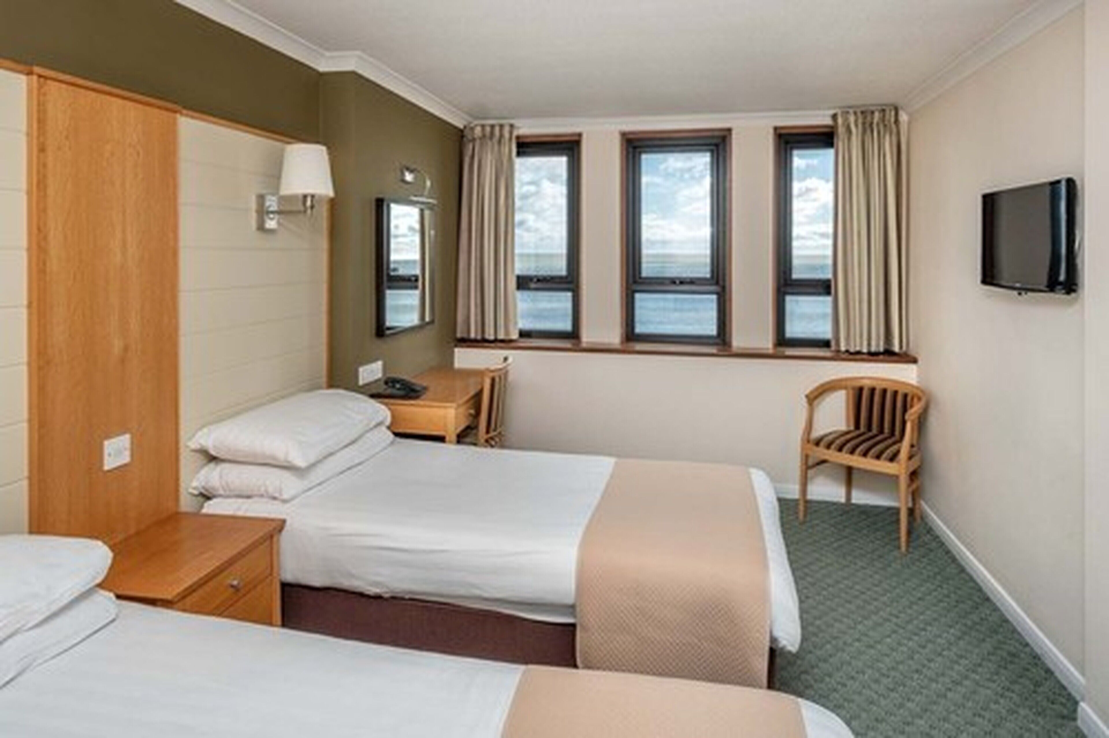 A hotel room with two beds, a desk, a chair, a TV, and a view of the sea through large windows.