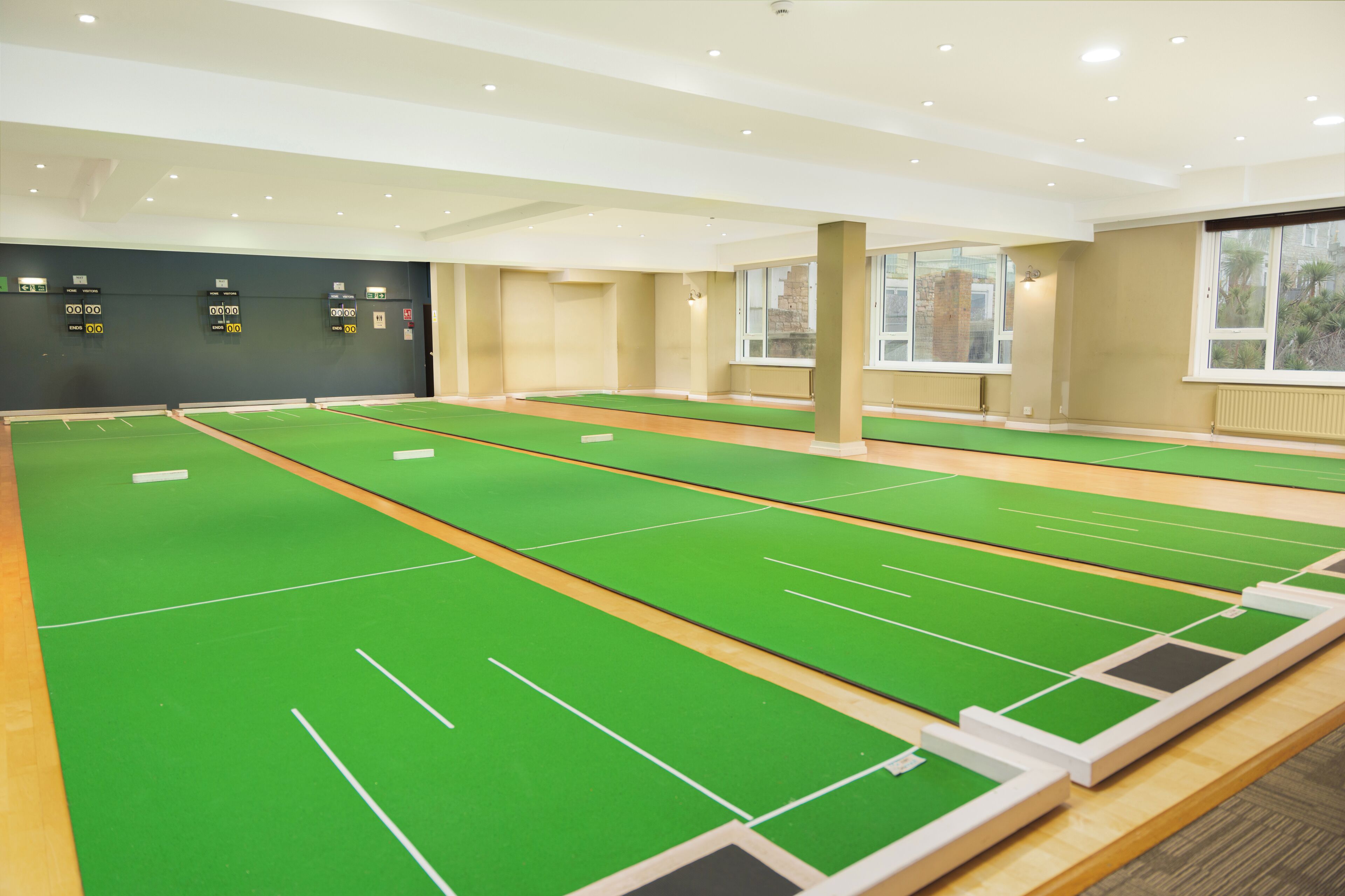 Una amplia cancha de shuffleboard cubierta con varias pistas, una pared negra con marcadores de puntuación y grandes ventanales que proporcionan luz natural.