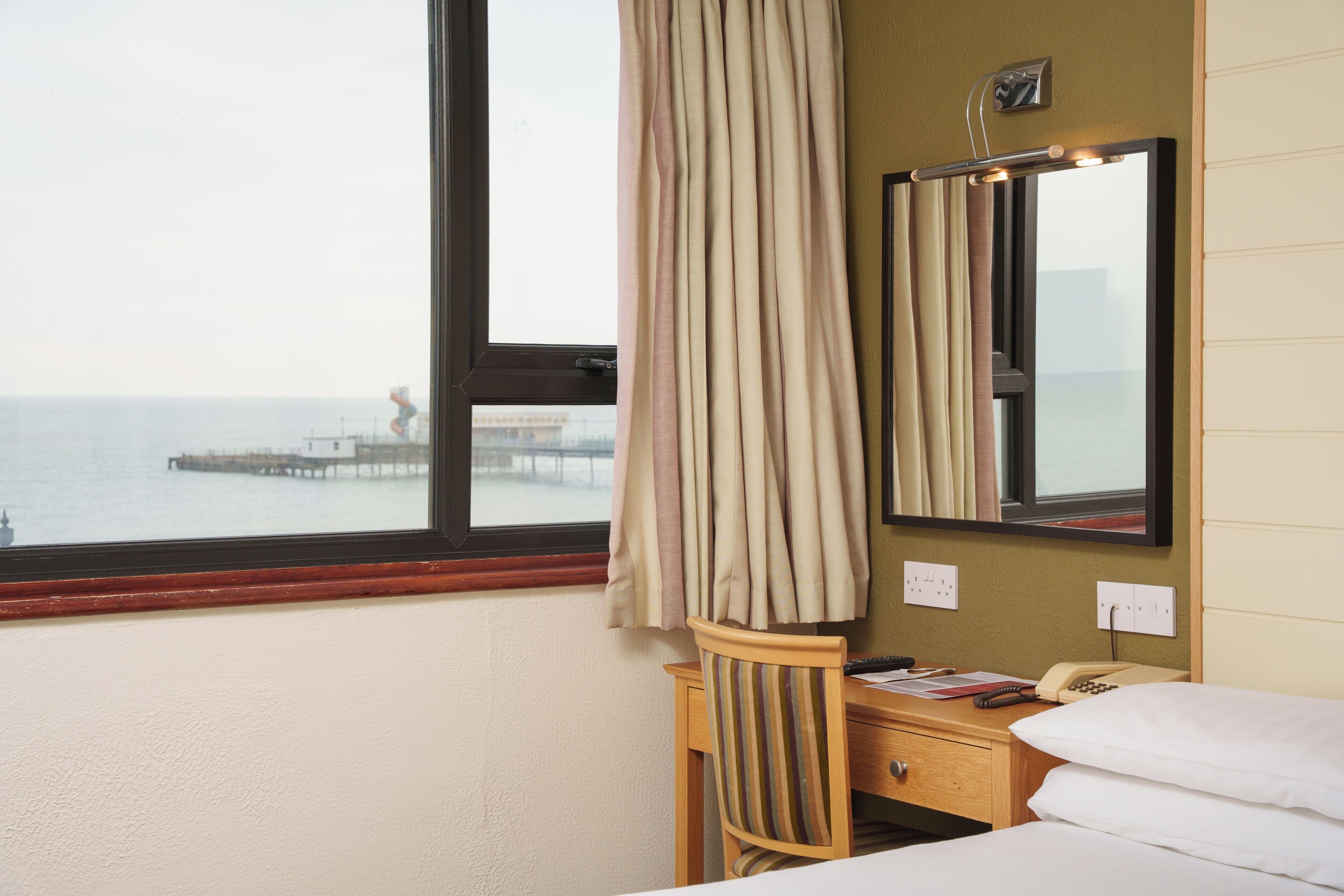 A hotel room with a bed, a wooden nightstand, a mirror, and a view of the sea through a window.