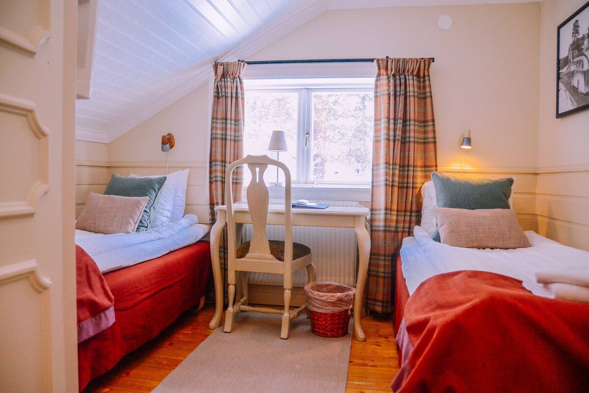 Ein kleines Zimmer mit zwei Betten, einem Sessel, einem Tisch und einem Fenster mit Vorhängen.