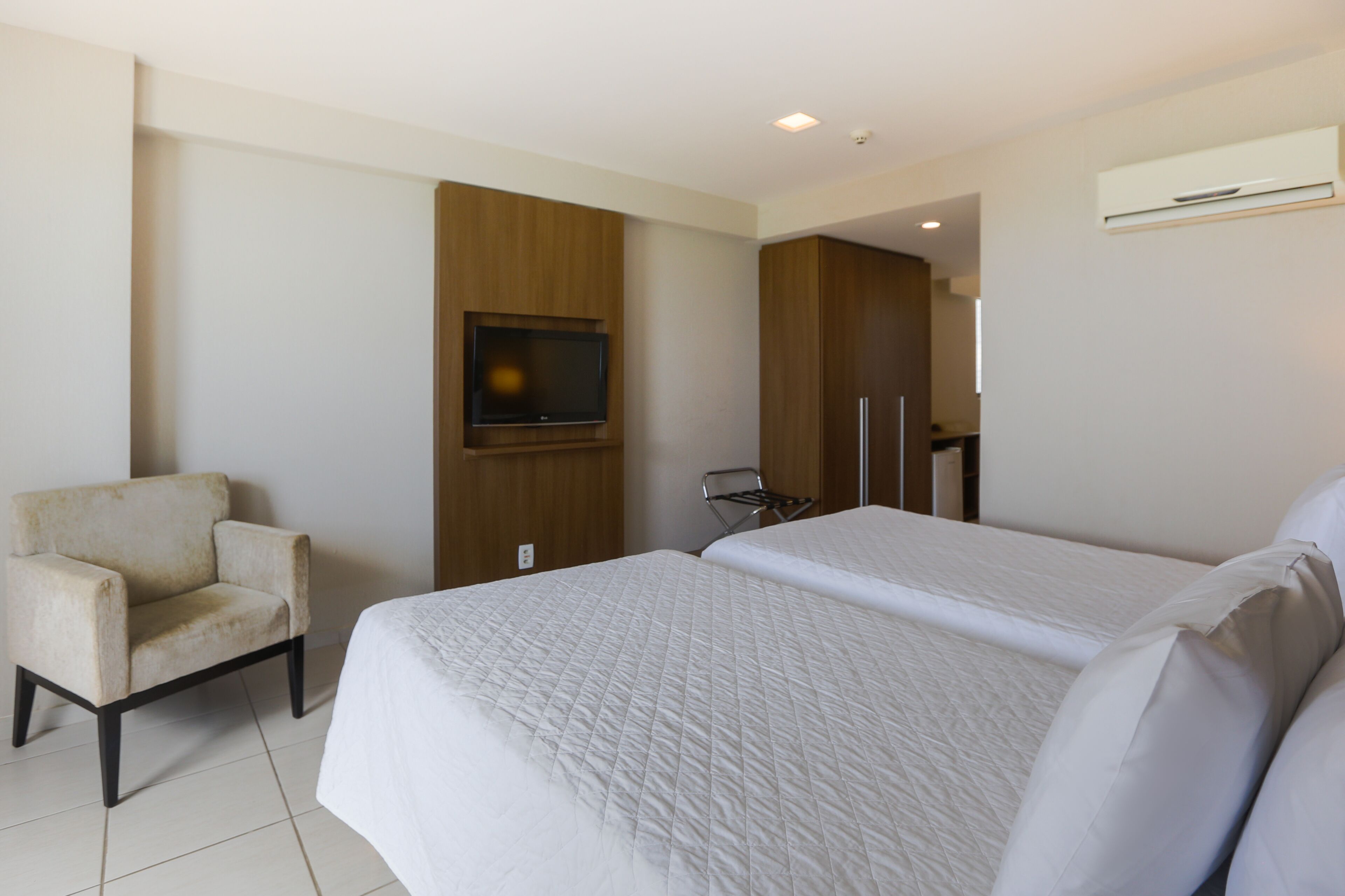A hotel room with a large bed, a chair, a television mounted on the wall, and an air conditioning unit.