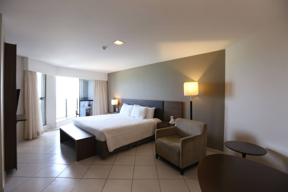 A hotel room with a large bed, a bedside table, a chair, a small table, and a lamp.