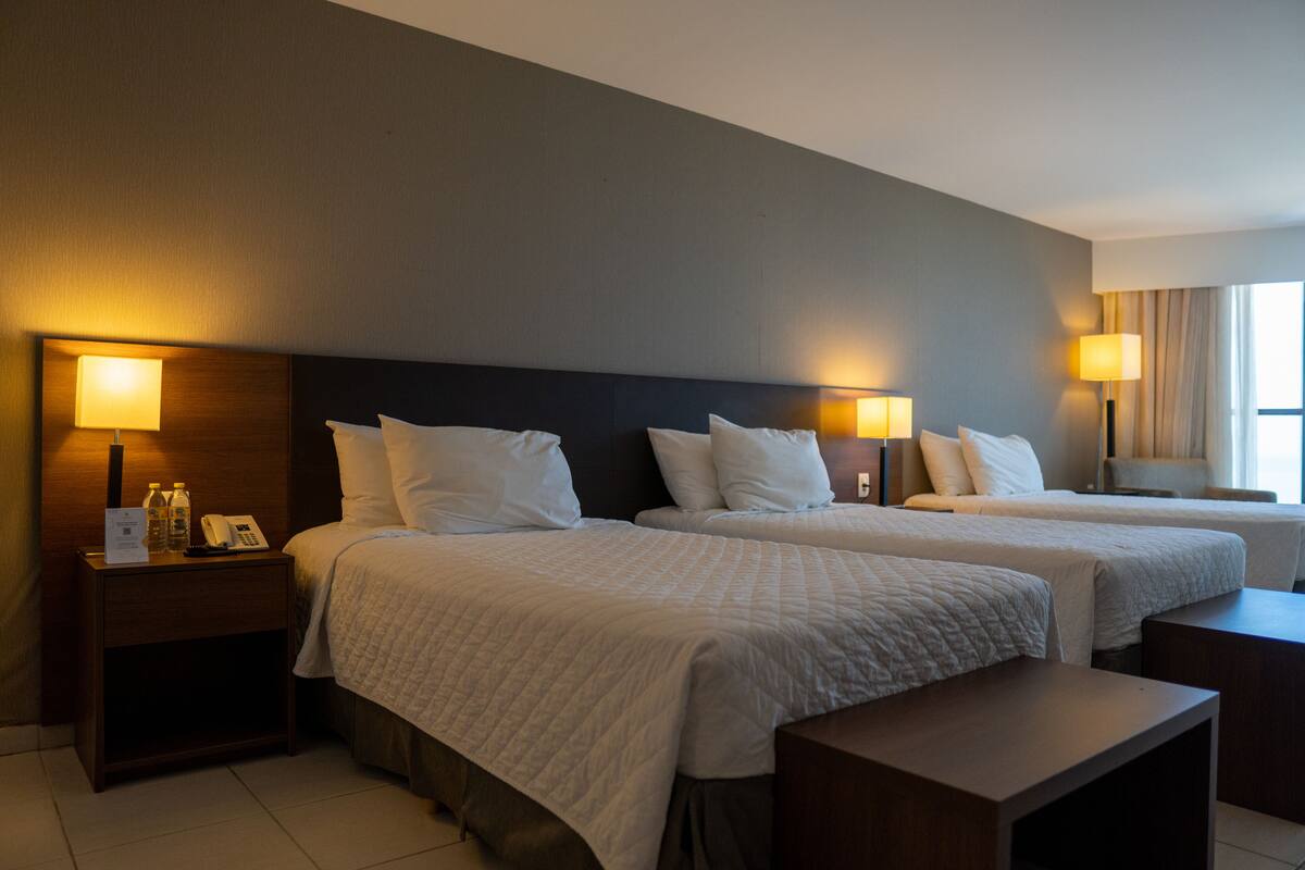 A hotel room with two beds, each with a headboard and bedside lamps. There is a wooden nightstand with a phone and a bottle of water.