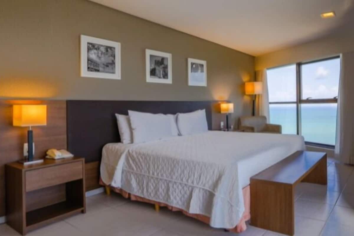 A hotel room with a large bed, bedside lamps, a nightstand, a wooden bench, and a large window with a view.
