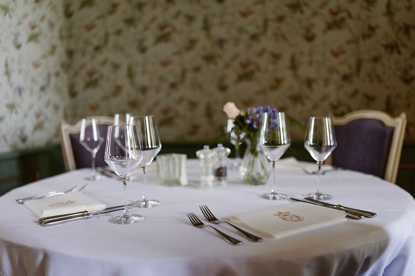 A round dining table set with multiple wine glasses, cutlery, and a folded menu.