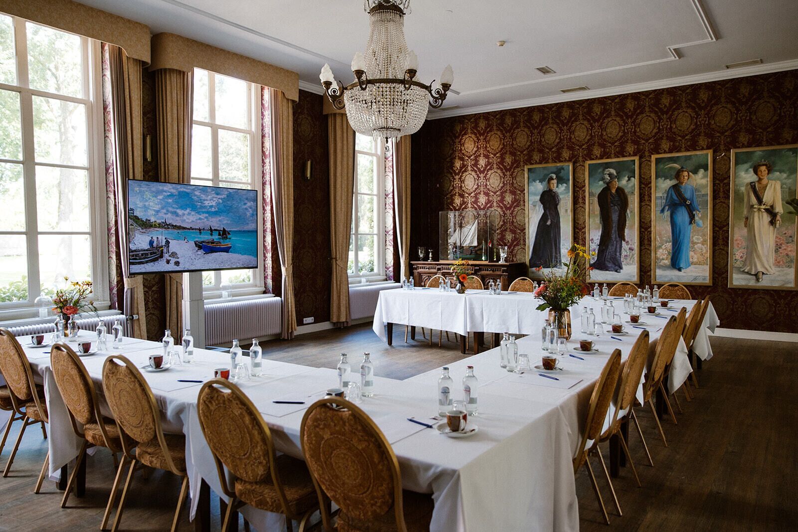 Un comedor formal con mesas largas, sillas, un candelabro y ventanas amplias con cortinas.