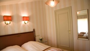 Habitación de hotel con cama, dos lámparas de pared, una mesita de noche con un florero y una puerta con un espejo que refleja la cama.