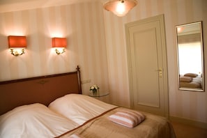 Une chambre d’hôtel avec un lit, deux lampes fixées au mur, une petite table de chevet ornée d’un vase de fleurs et une porte dotée d’un miroir qui reflète le lit.