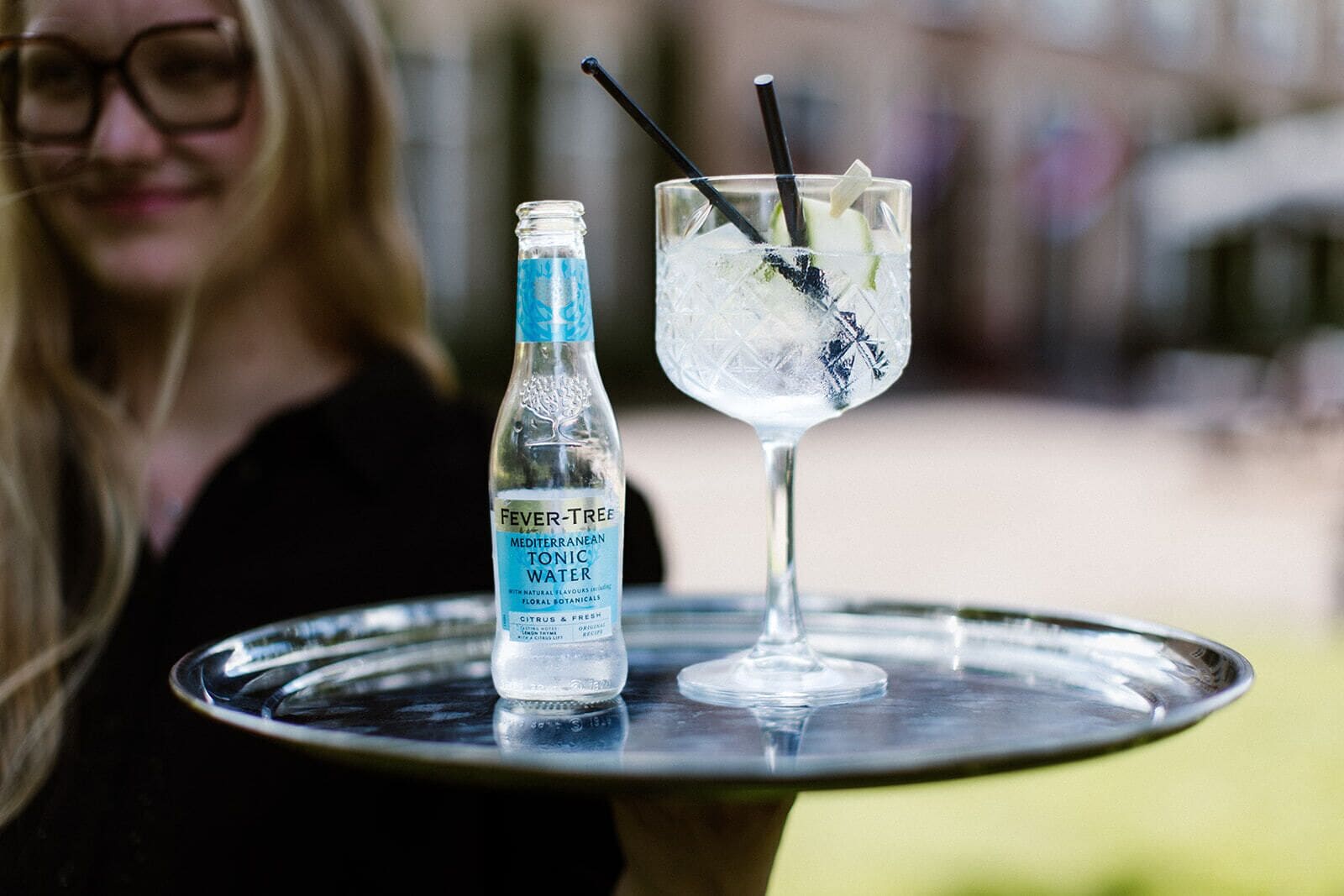 Una persona sosteniendo una bandeja con una botella de Fever-Tree Tónica Mediterránea y un vaso con una bebida.