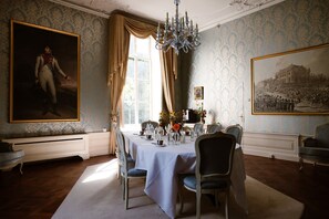Un comedor formal con una mesa dispuesta para cuatro, rodeada de sillas ornamentadas, una araña y dos cuadros grandes enmarcados.