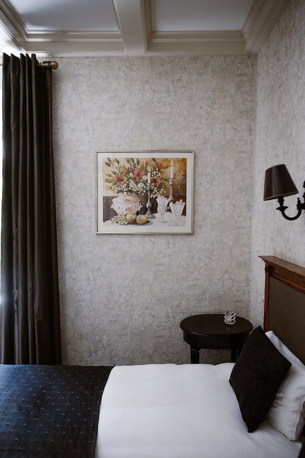 A hotel room with a bed, a small round table, a wall-mounted lamp, and a framed floral painting.