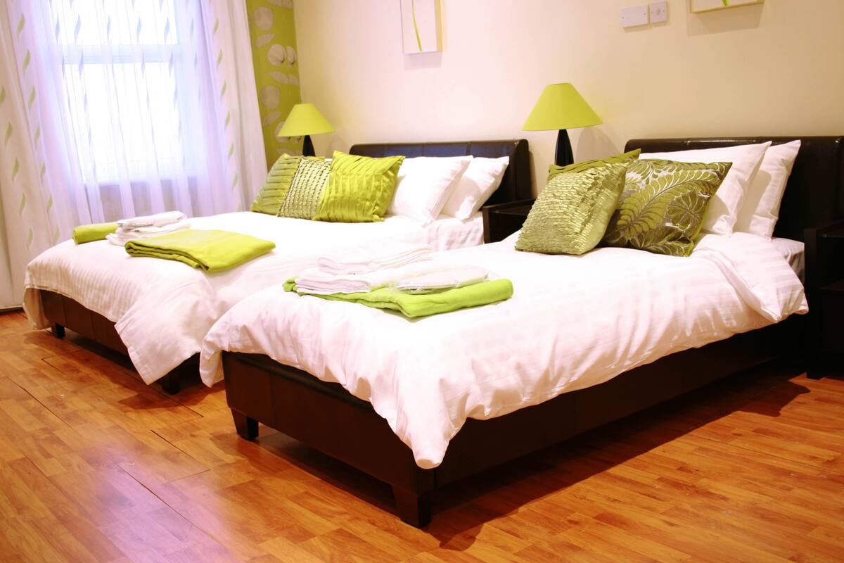 A double bed with a white and green bedspread, two green pillows, and a green blanket folded on the bed.