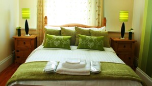 A neatly made bed with green pillows and a green blanket, two bedside lamps, and a window with curtains. - Hotel Makedonia (London)