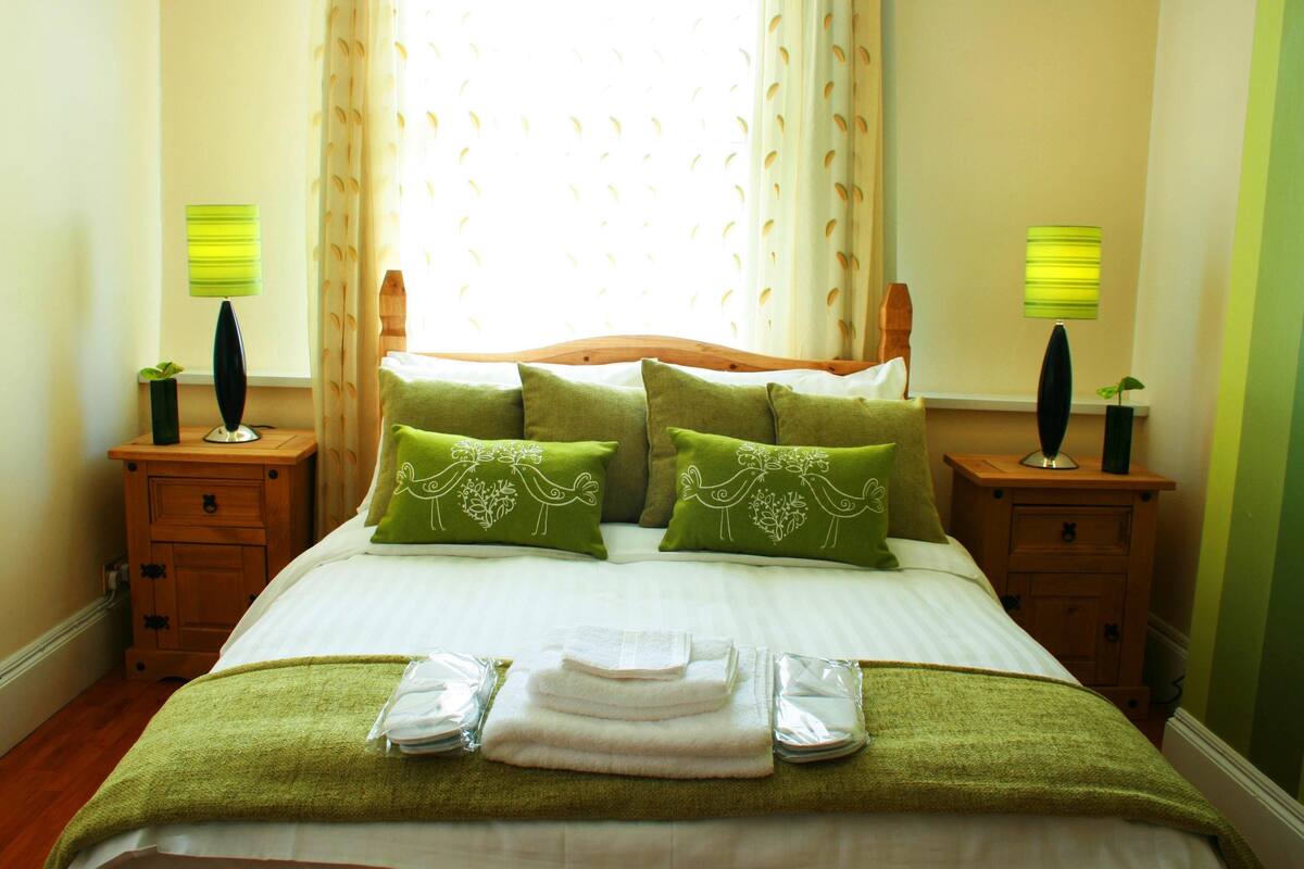 A neatly made bed with green pillows and a green blanket, two bedside lamps, and a window with curtains.