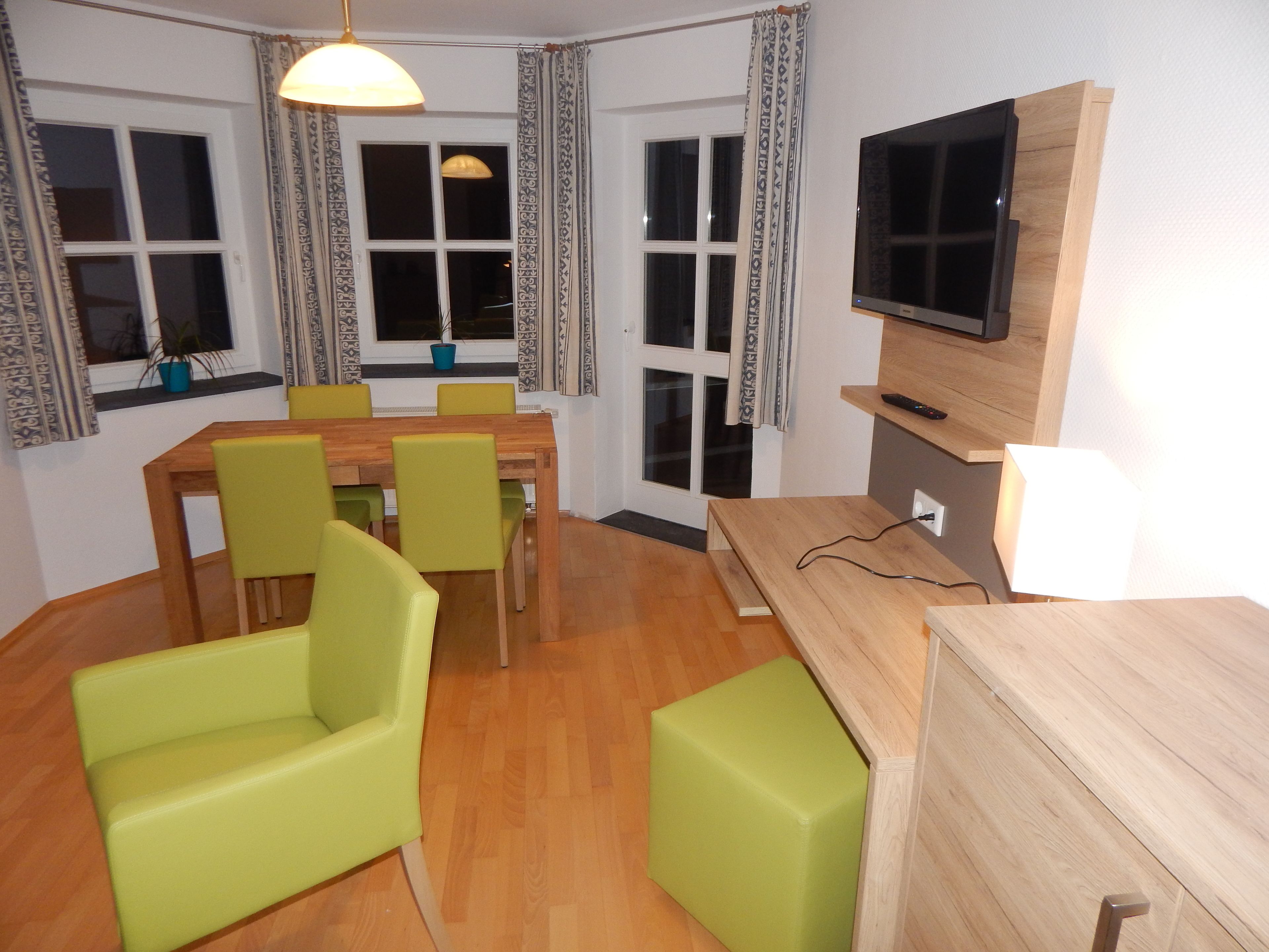 A room with a wooden dining table, four green chairs, a TV on a wooden stand, and a window with patterned curtains.