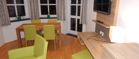 A room with a wooden dining table, four green chairs, a TV on a wooden stand, and a window with patterned curtains.