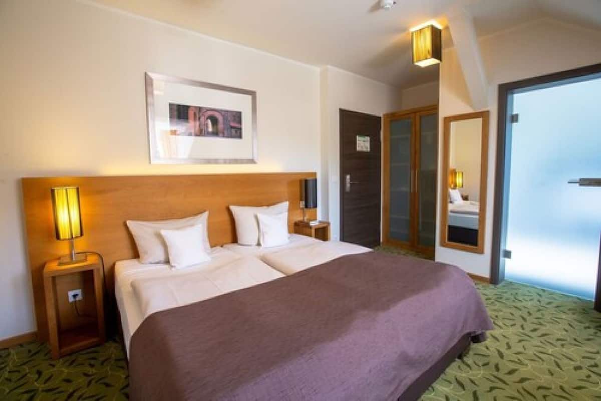 A hotel room with a large bed, a bedside table with a lamp, a framed picture on the wall, and a door leading to another room.