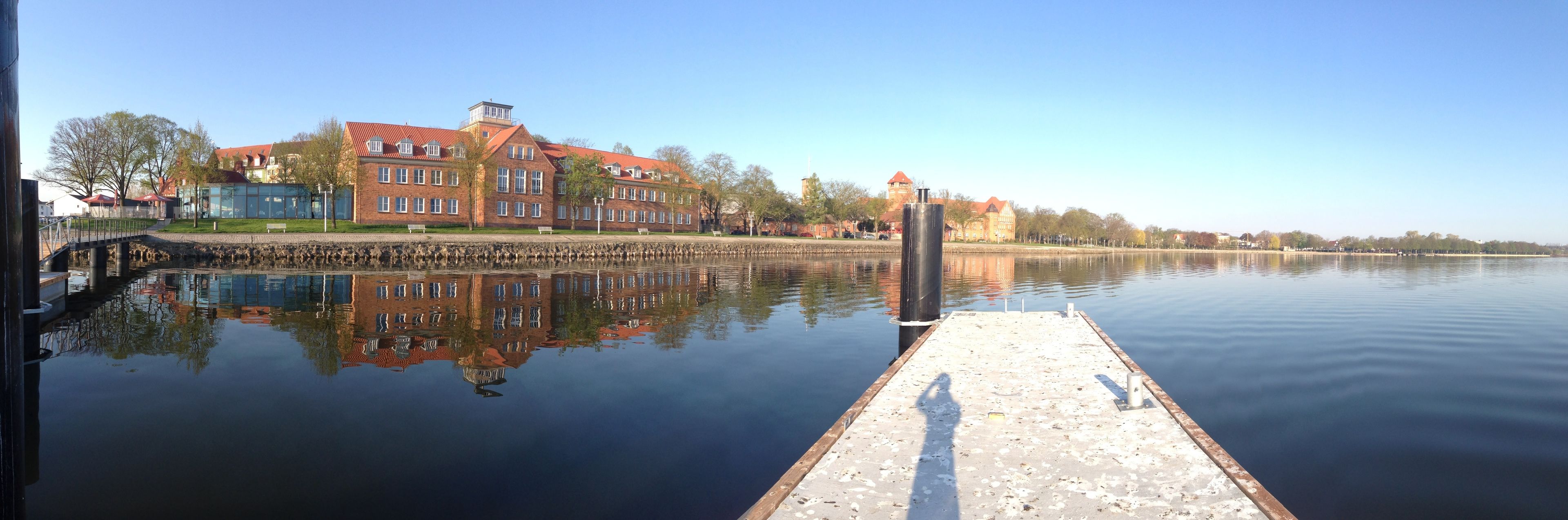 A calm lake with a wooden dock extending into the water, surrounded by buildings with red roofs and a clear sky.