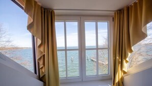 Quarto com uma janela ampla com vista para um corpo d'água, cortinas e decoração em tons de branco.