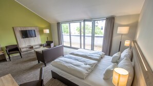 Quarto de hotel com uma cama grande, uma escrivaninha, uma cadeira, televisão fixada na parede e vista para o exterior pelas janelas.