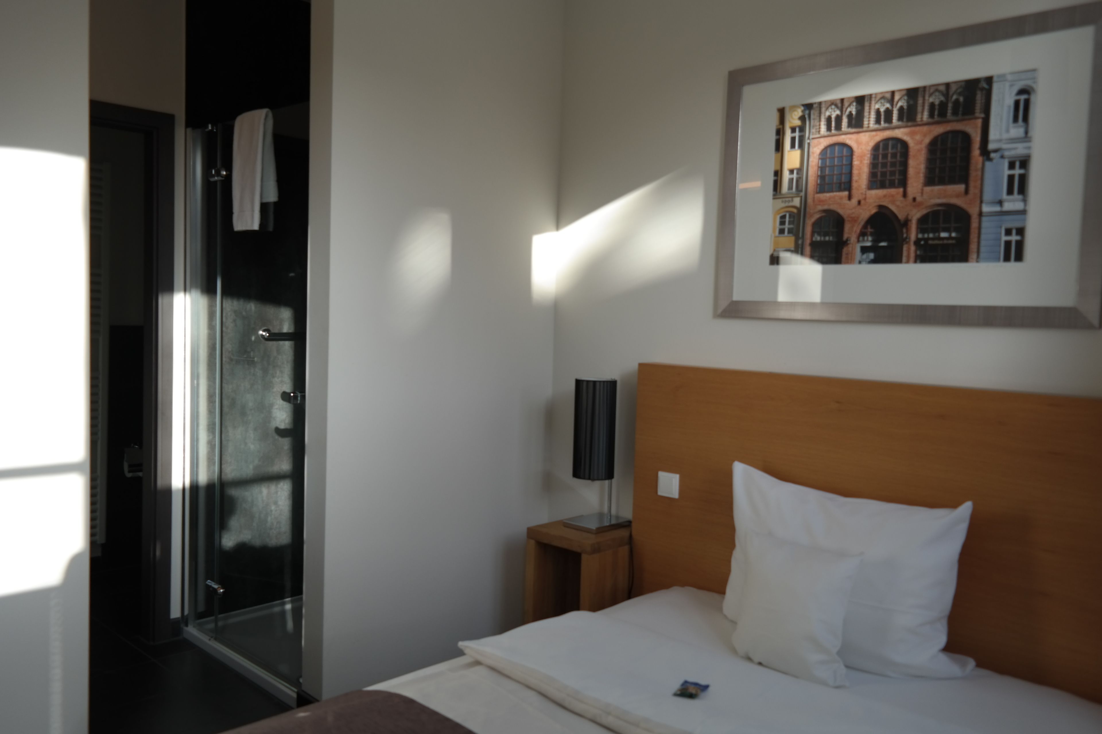 Una habitación de hotel con una cama, una mesita de noche, un cuadro enmarcado en la pared y una puerta que da a un baño.