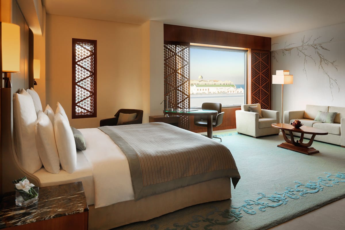A hotel room with a bed, two armchairs, a sofa, a dining table, and a view of a building through a window.